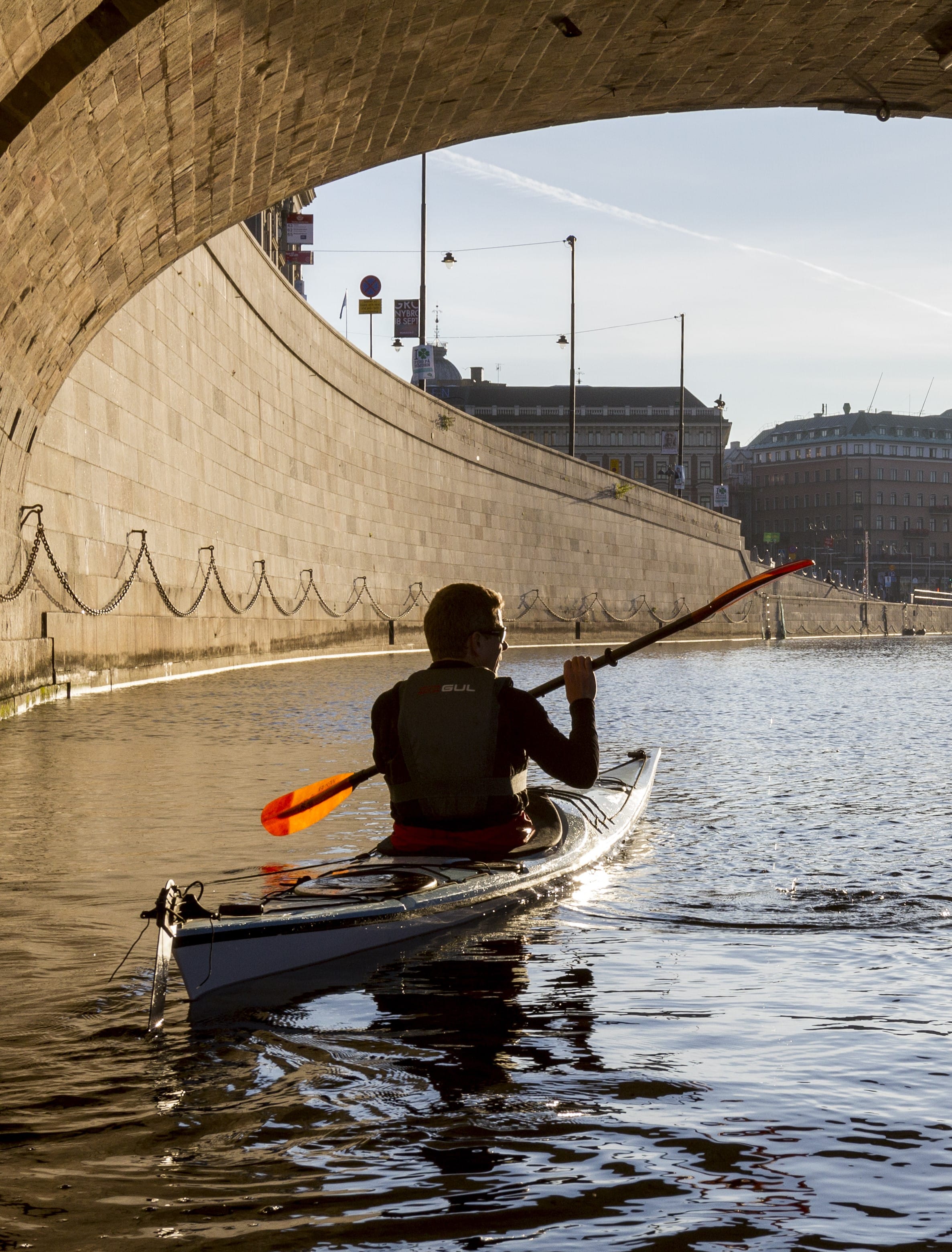 A person kayaking on a calm waterway beneath a bridge, with city buildings in the background and sunlight reflecting on the water.