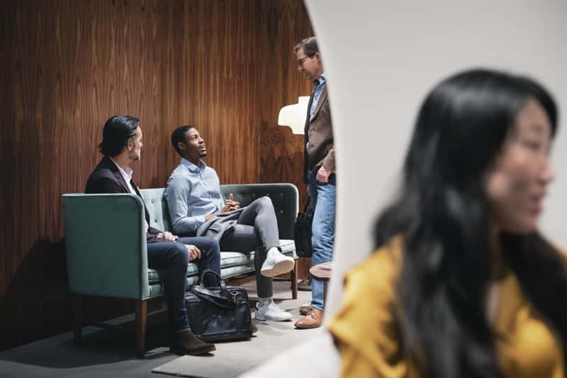 Four people in business attire conversing in a stylish lounge with wood paneling and a teal sofa.
