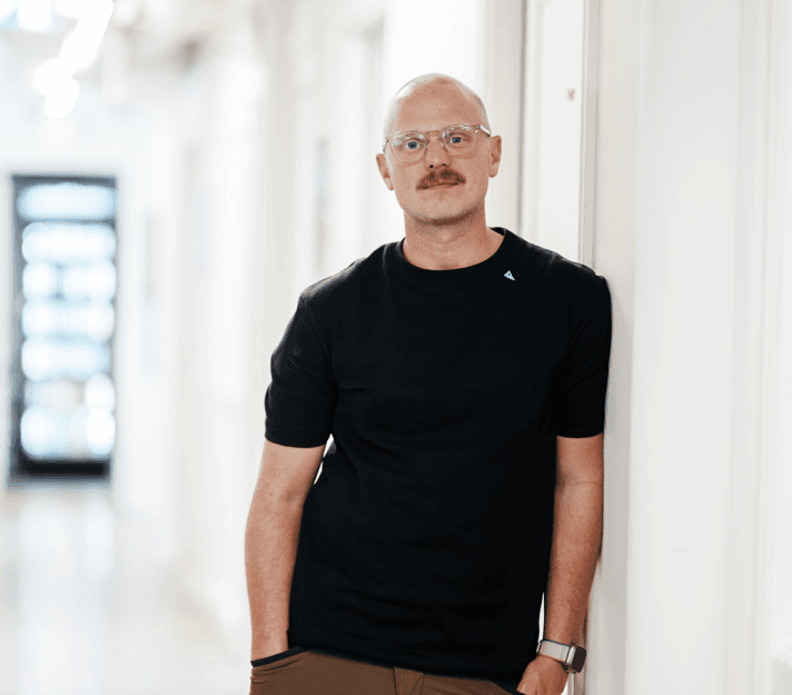 Mange Villson Mange Villson looking amicably at the spectator and leaning against a white wall, wearing a black t-shirt. He is a middle-aged man with a shaved head, a moustasche and transparent glasses.