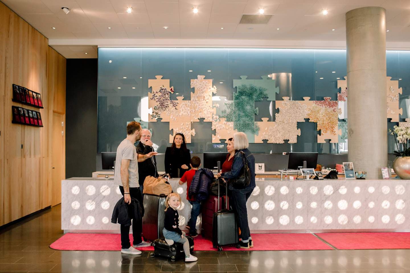A family with children checks in at a modern hotel reception. Several adults stand by the counter with luggage while a girl sits smiling on a suitcase.