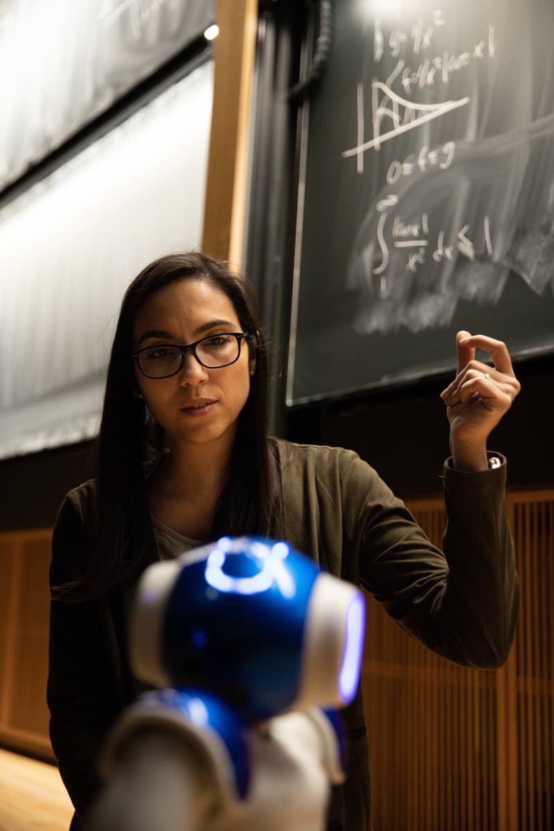 Woman wearing glasses stands by a chalkboard with math formulas, gesturing while a small blue robot is visible in the foreground.