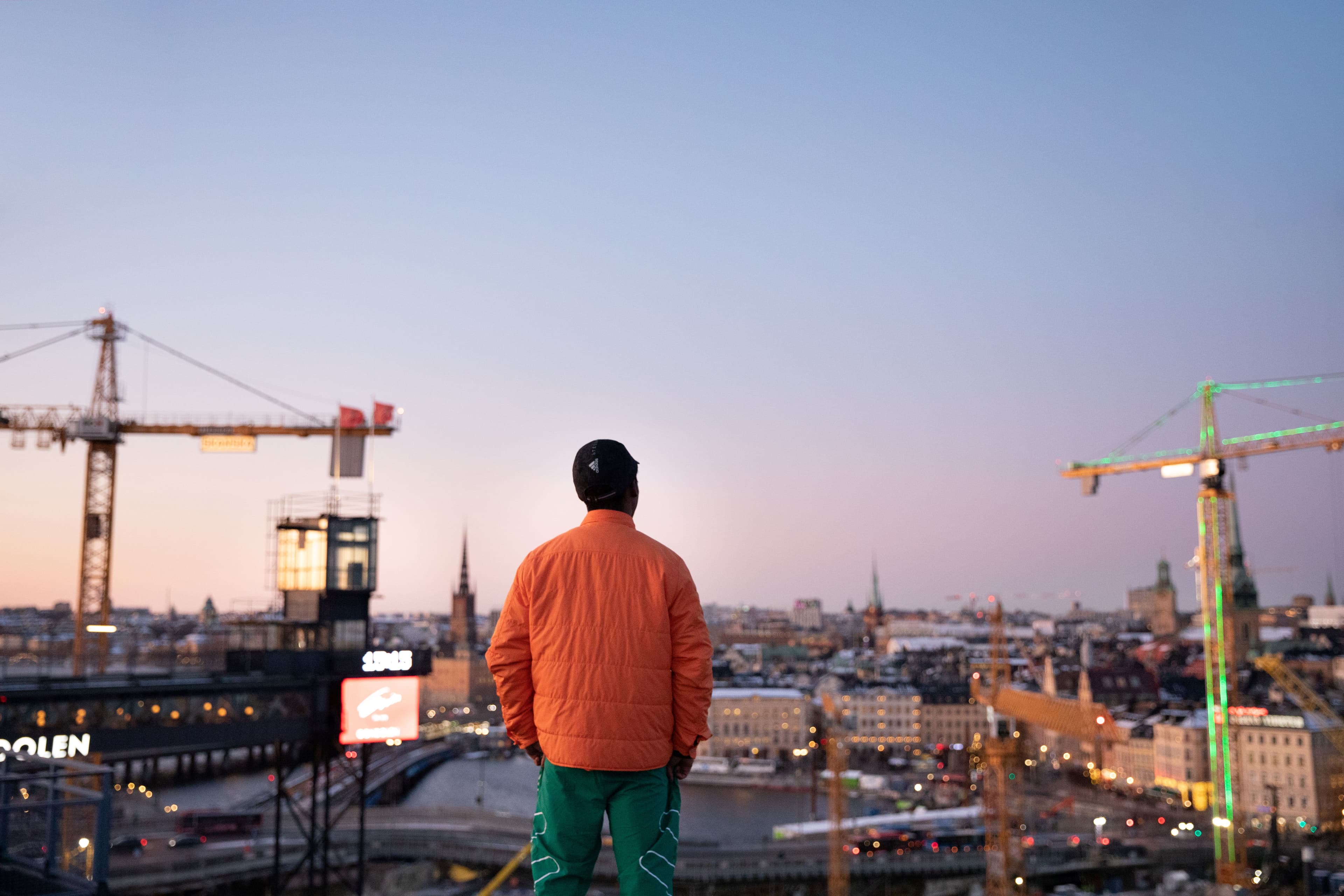 Person in an orange jacket stands with their back to the camera, overlooking Stockholm’s skyline and construction cranes at dusk.