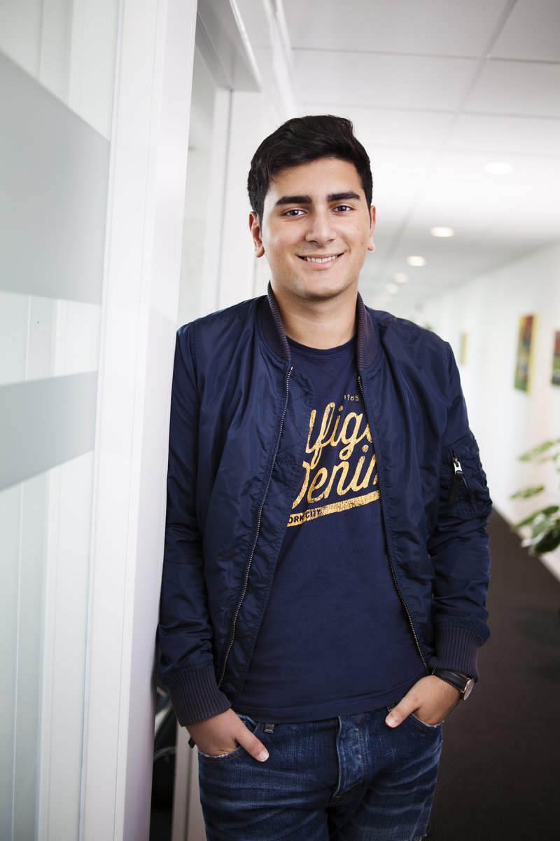 En ung man står lutad mot en vägg i en ljus kontors- eller skolmiljö och ler mot kameran. Han bär jacka och jeans med händerna i fickorna. Bilden förmedlar en avslappnad och vänlig stämning.